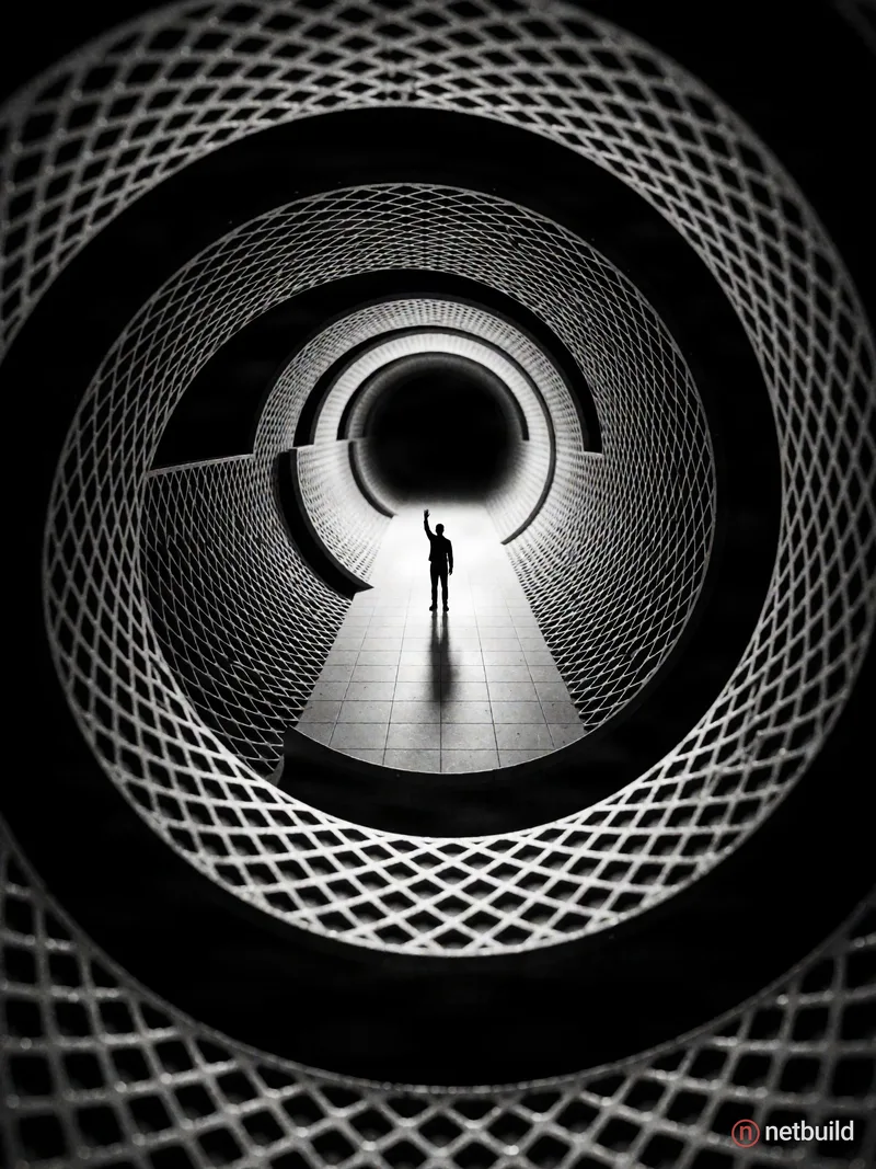 A high-contrast black-and-white photograph of a surreal circular tunnel made of curved architectural grids and mesh. At the exact center, a tiny silho...