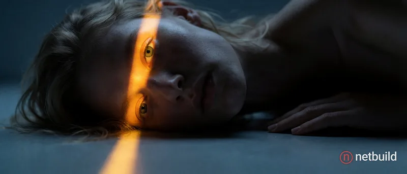 Cinematic close-up portrait of a fair-skinned person resting sideways on a smooth cool-gray surface. A stark, narrow beam of vivid golden-orange light...