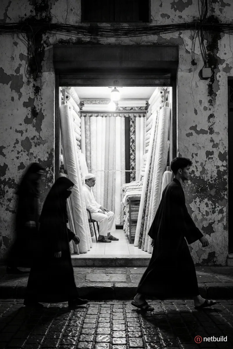 High-contrast black and white street photography. A brightly lit fabric shop framed by heavily peeling exterior walls. Inside the open storefront, a m...
